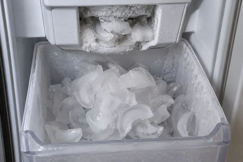 Frost-coated ice bin holding thin, hollow crescent cubes under an ice outlet jammed with frost